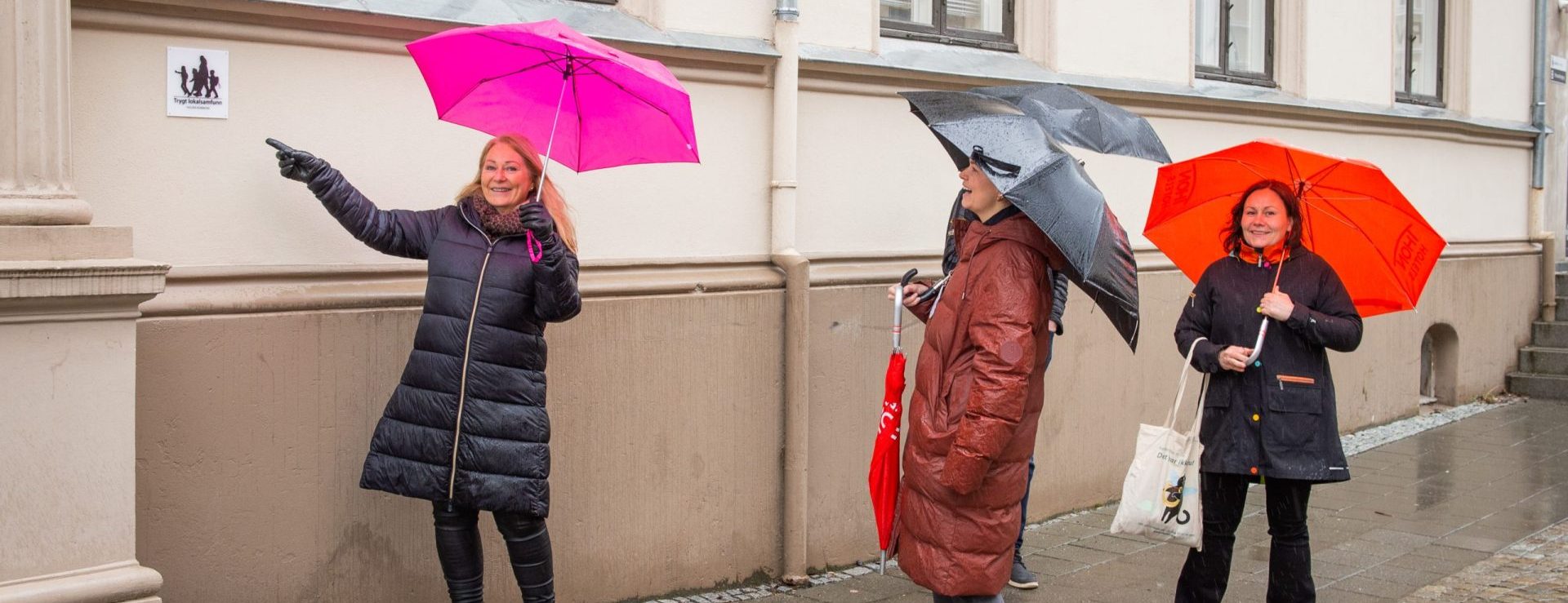 Tre kvinner fra Trygge lokalsamfunn med paraplyer ser mot et skilt. Den ene damen peker på skiltet. Foto.