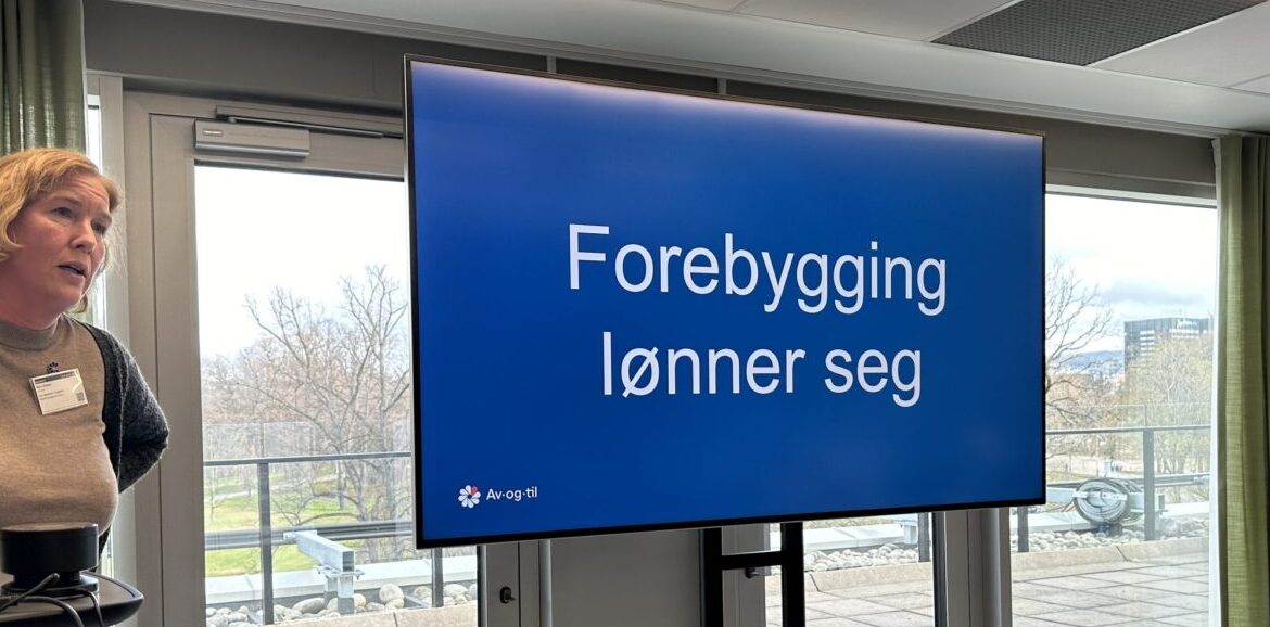 En dame står og snakker. Ved siden av er en skjerm der det står skrevet: Forebygging lønner seg. Foto.